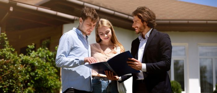 Millennial couple and realtor looking through house ownership documents near residential building outdoors, panorama